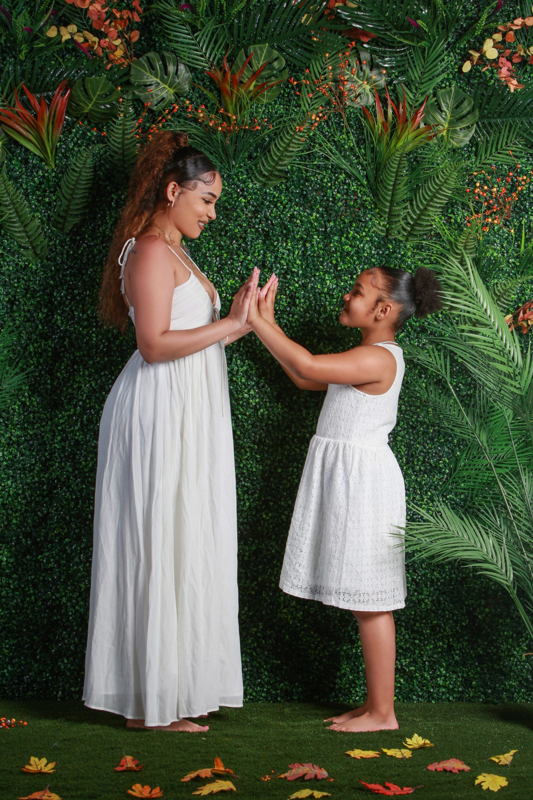 Mother and daughter in white dresses pressing palms together in front of a greenery wall